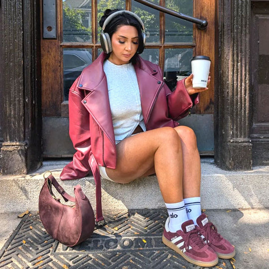 Person sitting in a doorway next to a Nora Shoulder Bag with headphones on holding a hot drink up